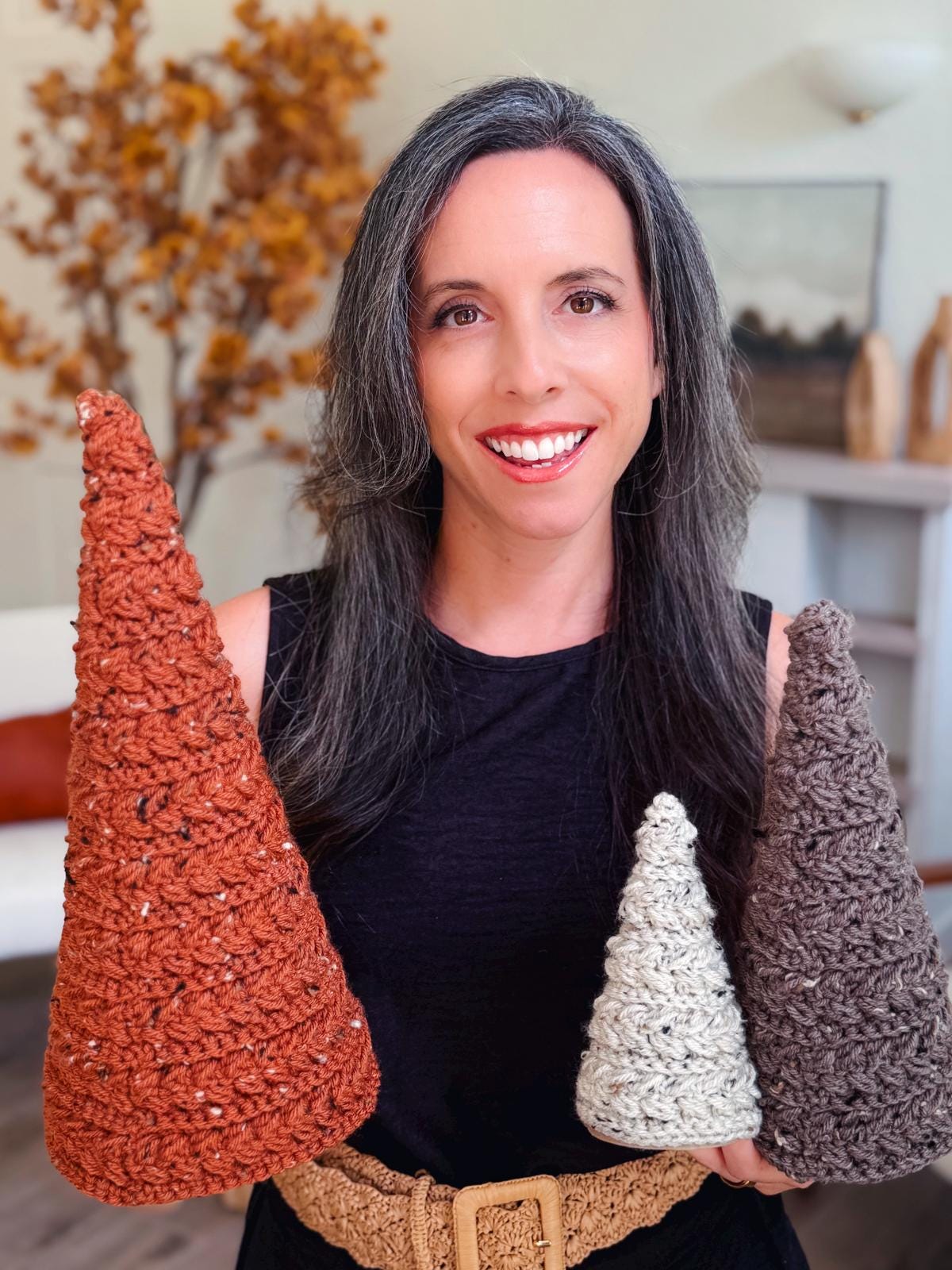 A woman with long dark and gray hair smiles while holding three crocheted cone-shaped objects—perfect as a handmade gift—standing indoors with autumn decor in the background.