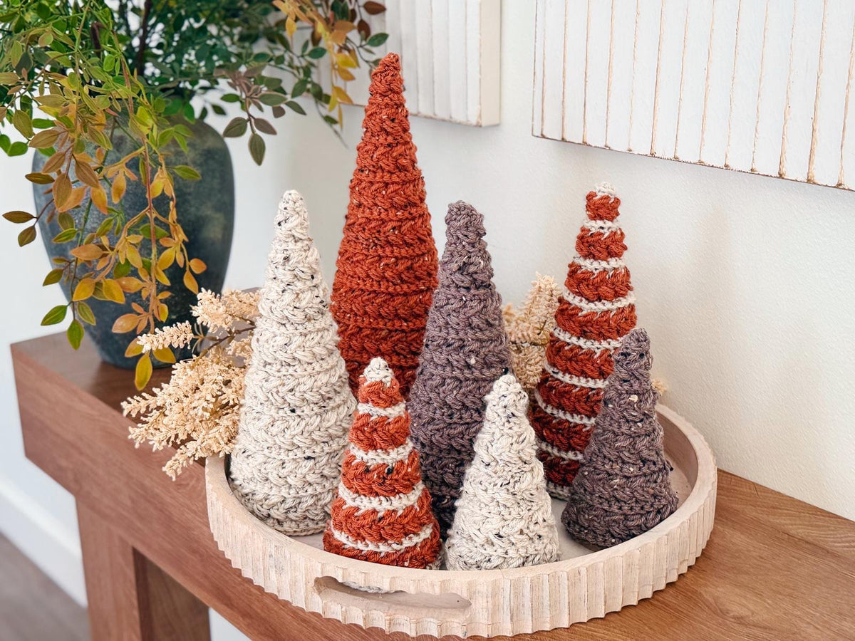 A wooden tray displays seven crocheted decorative trees in various earth tones, arranged with dried foliage on a wooden table beside a vase of greenery—perfect as a charming handmade gift.