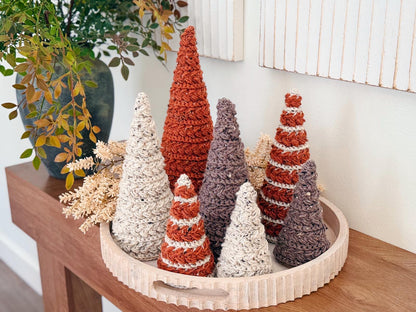 A wooden tray displays seven crocheted decorative trees in various earth tones, arranged with dried foliage on a wooden table beside a vase of greenery—perfect as a charming handmade gift.