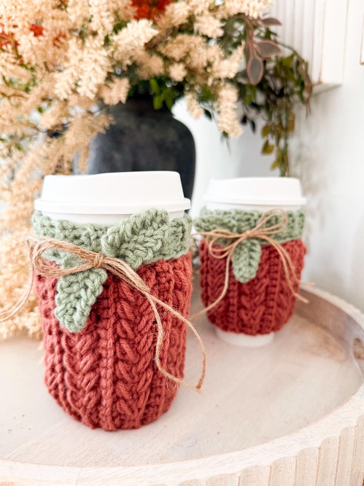 Two takeaway coffee cups wrapped in crochet, two-tone cozies with twine bows, placed on a white tray—perfect as a handmade gift, with a blurred floral arrangement in the background.