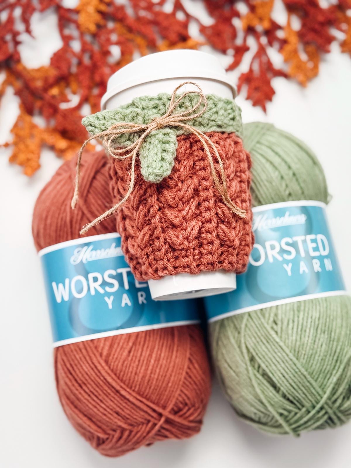 A coffee cup with a crocheted pumpkin-shaped cozy, perfect as a handmade gift, rests on two skeins of worsted yarn—one rust-colored and one green—with autumn leaves in the background.