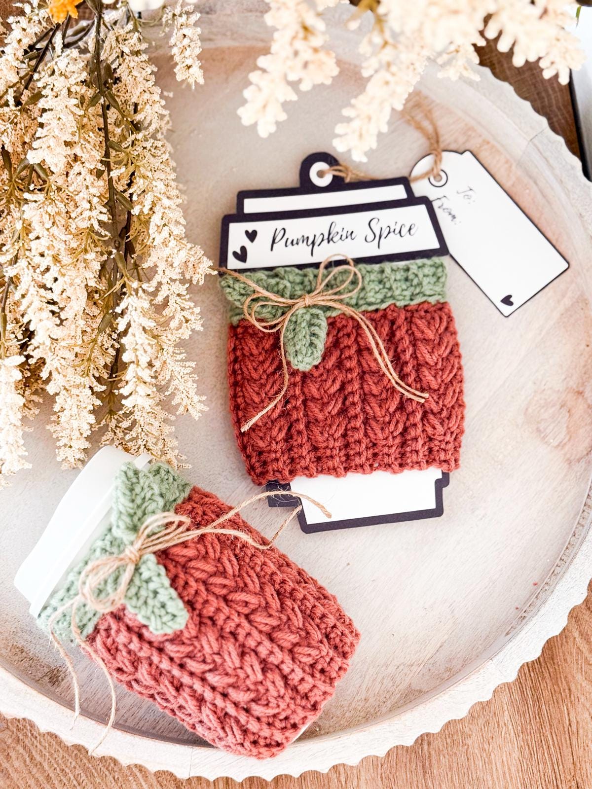 Two takeaway coffee cups with handmade crochet sleeves in autumn colors sit on a tray next to decorative foliage. A tag reads "Pumpkin Spice." This cozy scene feels like the perfect inspiration for a handmade gift.