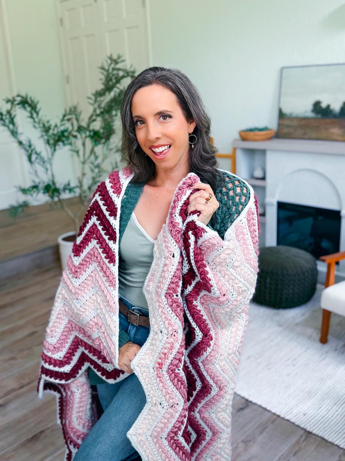 A woman stands indoors holding up a crocheted zigzag-patterned crochet blanket in shades of burgundy, pink, and white—a perfect handmade gift to cherish or share.