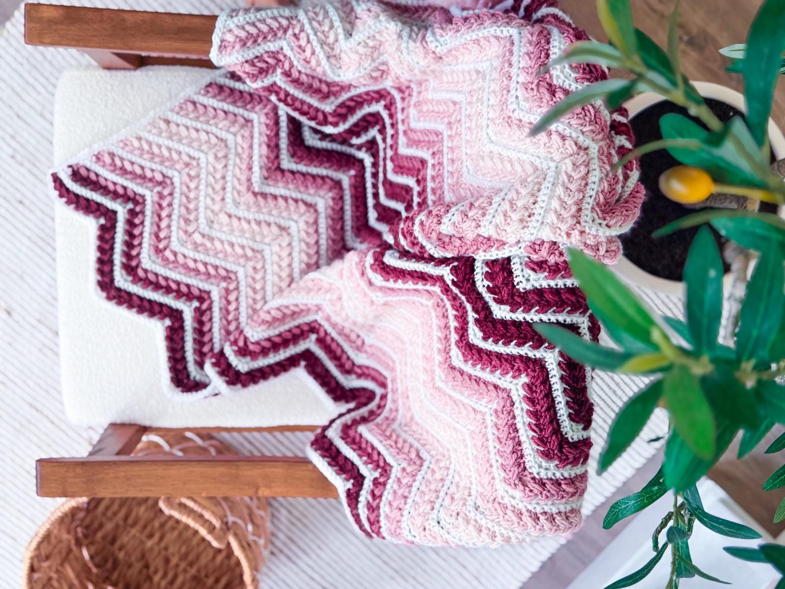 A pink, maroon, and white zigzag-patterned crocheted blanket draped over a wooden chair next to a green potted plant and a woven basket makes a perfect handmade gift.