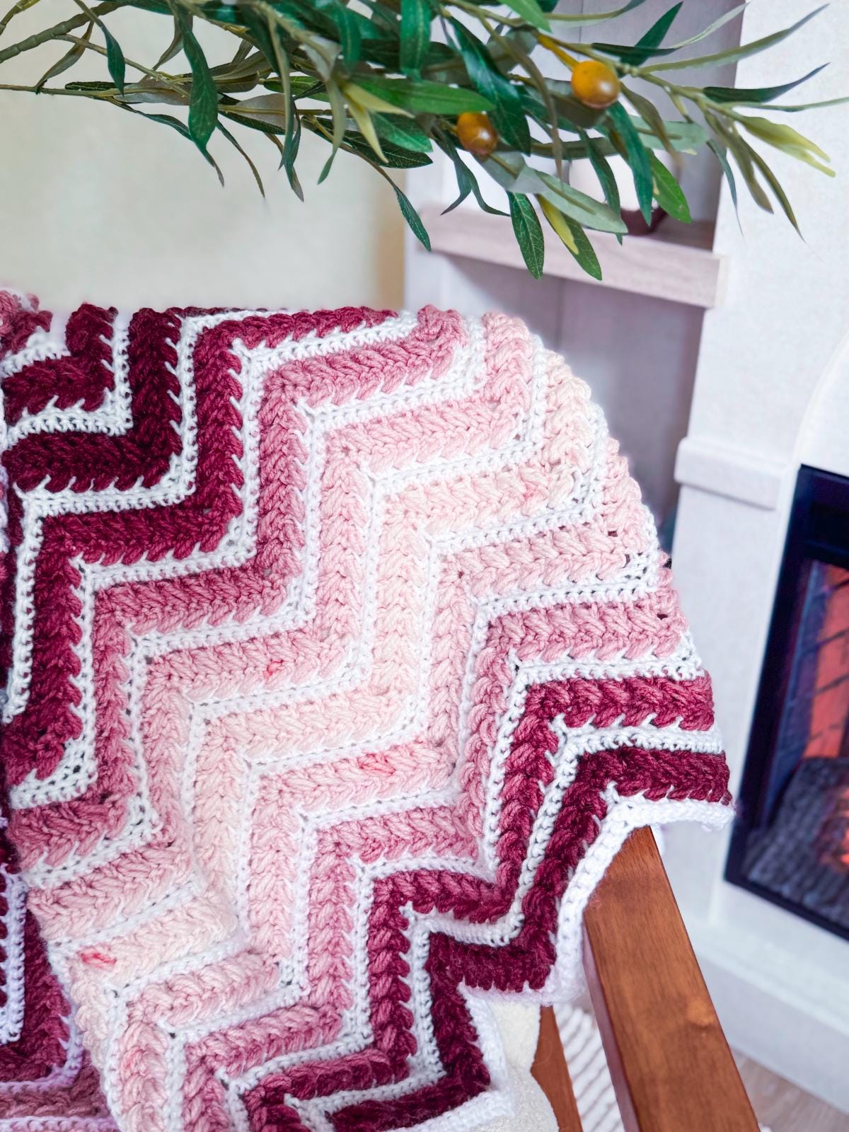 A crochet blanket with a chevron pattern in shades of burgundy, pink, and white—an ideal handmade gift—draped over a wooden chair near a fireplace and a leafy branch.