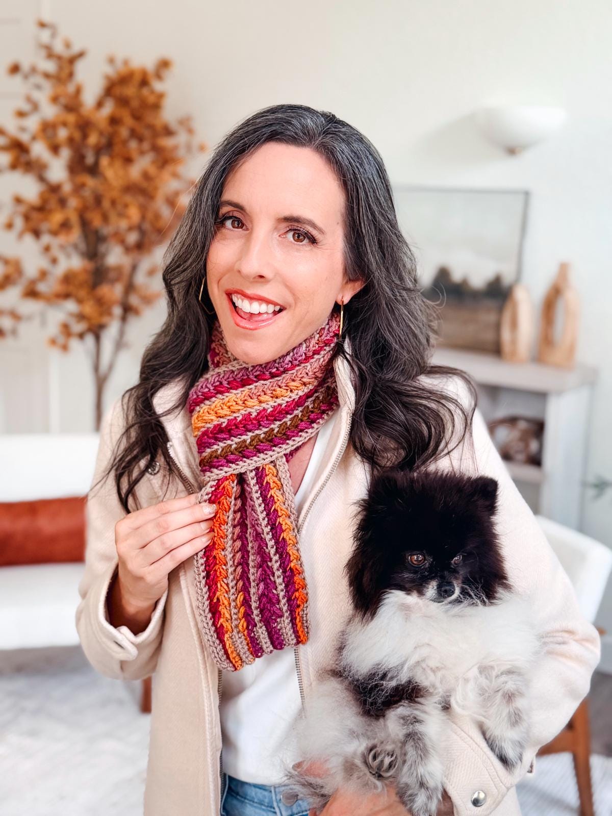 A woman with long dark hair and a streak of gray smiles at the camera, wearing a crochet scarf, light jacket, and white top—her style complemented by unique jewellery—in a bright room with autumn decor.