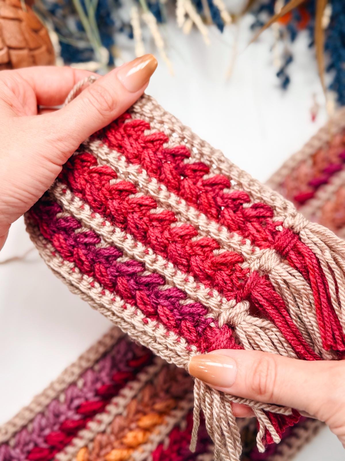 Hands holding a crochet piece with red and beige yarn, featuring a striped pattern and tassel fringes, with additional handmade crochet pieces and dried flowers softly arranged in the background.