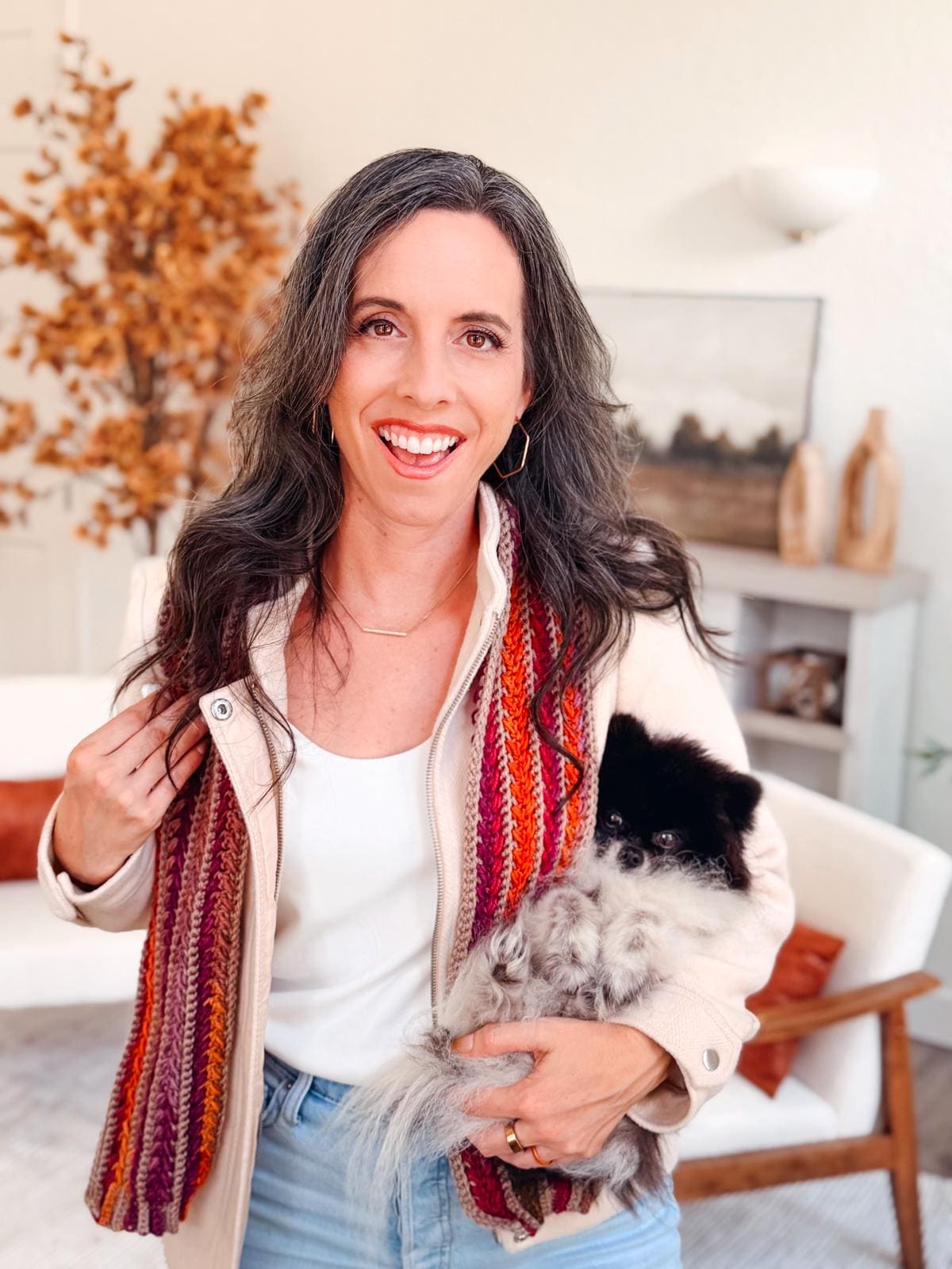 A woman with long dark hair and a streak of gray smiles at the camera, wearing a crochet scarf, light jacket, and white top—her style complemented by unique jewellery—in a bright room with autumn decor.