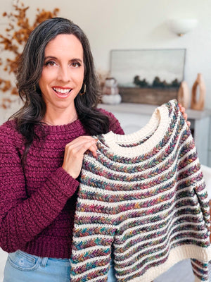 Multicolor striped crochet sweater held up by a smiling woman wearing a plum crochet version of the same sweater