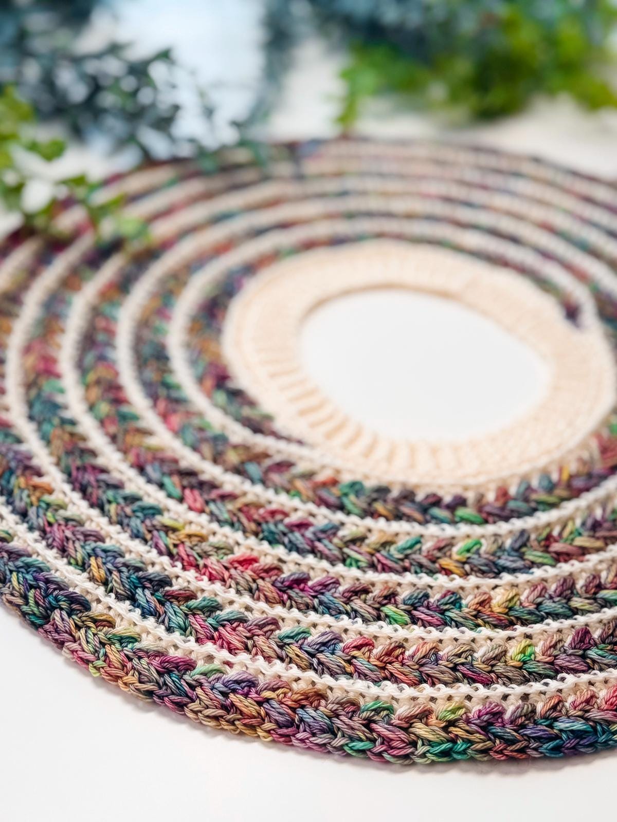 A round crocheted piece with a beige center and multicolored concentric rings, perfect as a handmade gift, is surrounded by green leafy branches on a white background.