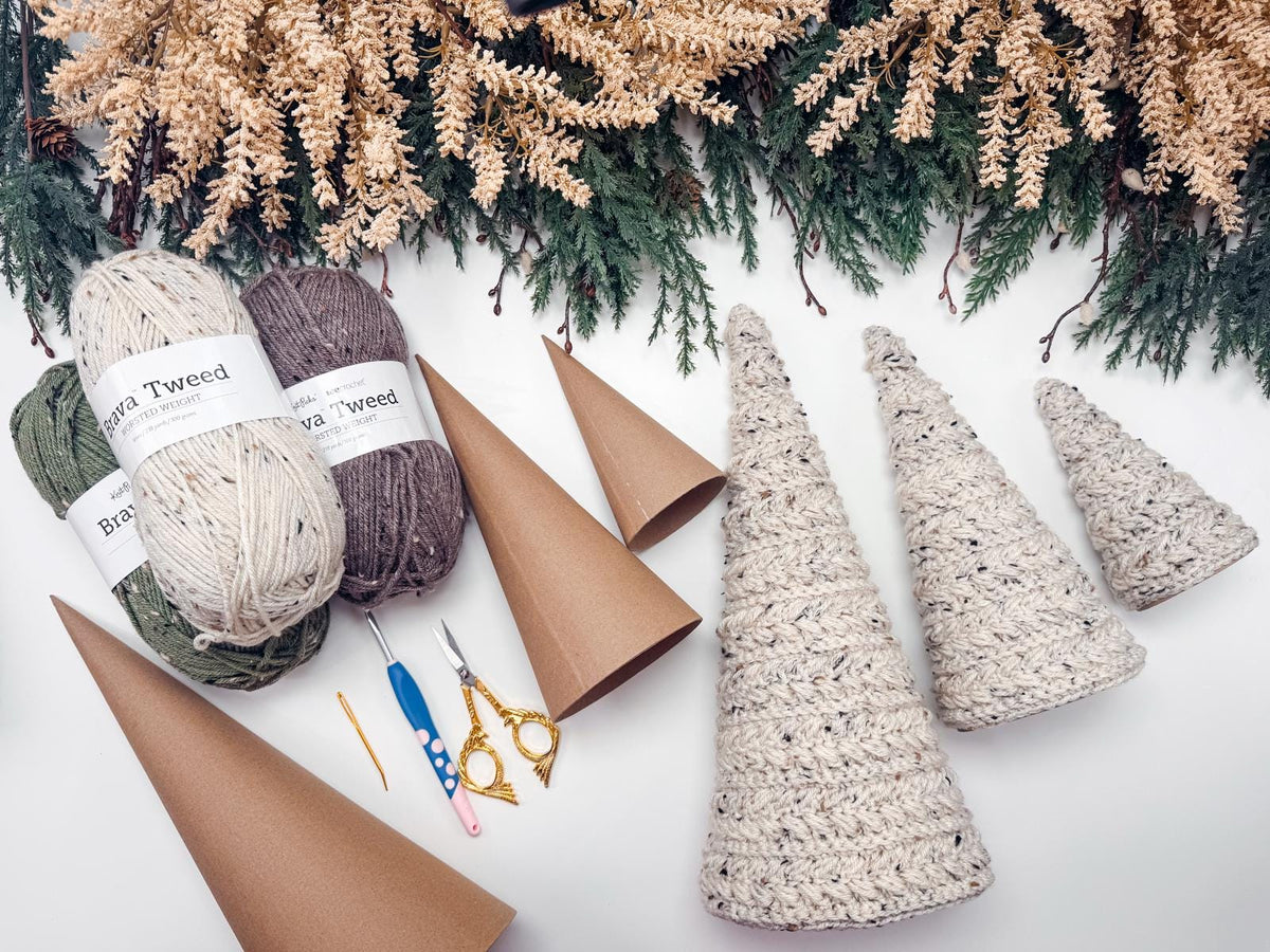 Yarn, crochet hooks, scissors, and needles are arranged with cardboard cones and finished crochet tree decorations on a white surface, perfect for a handmade gift, with pine branches at the top.