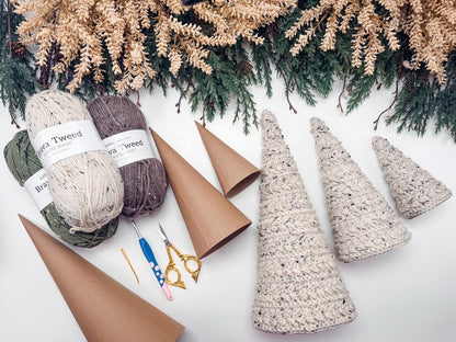 Yarn, crochet hooks, scissors, and needles are arranged with cardboard cones and finished crochet tree decorations on a white surface, perfect for a handmade gift, with pine branches at the top.
