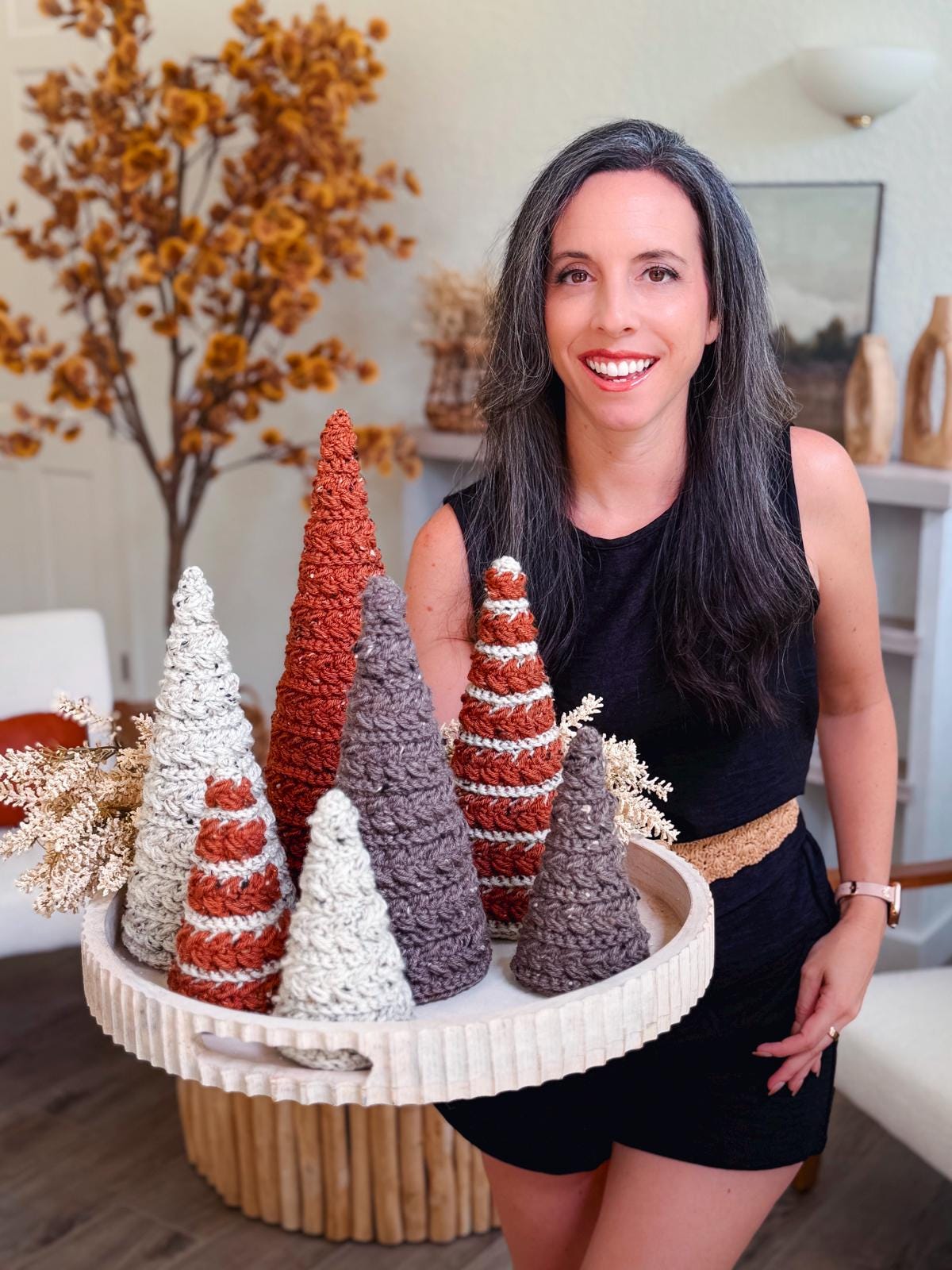 Crochet cone trees in rust, cream, and taupe, some striped, on a round tray held by a smiling woman in black