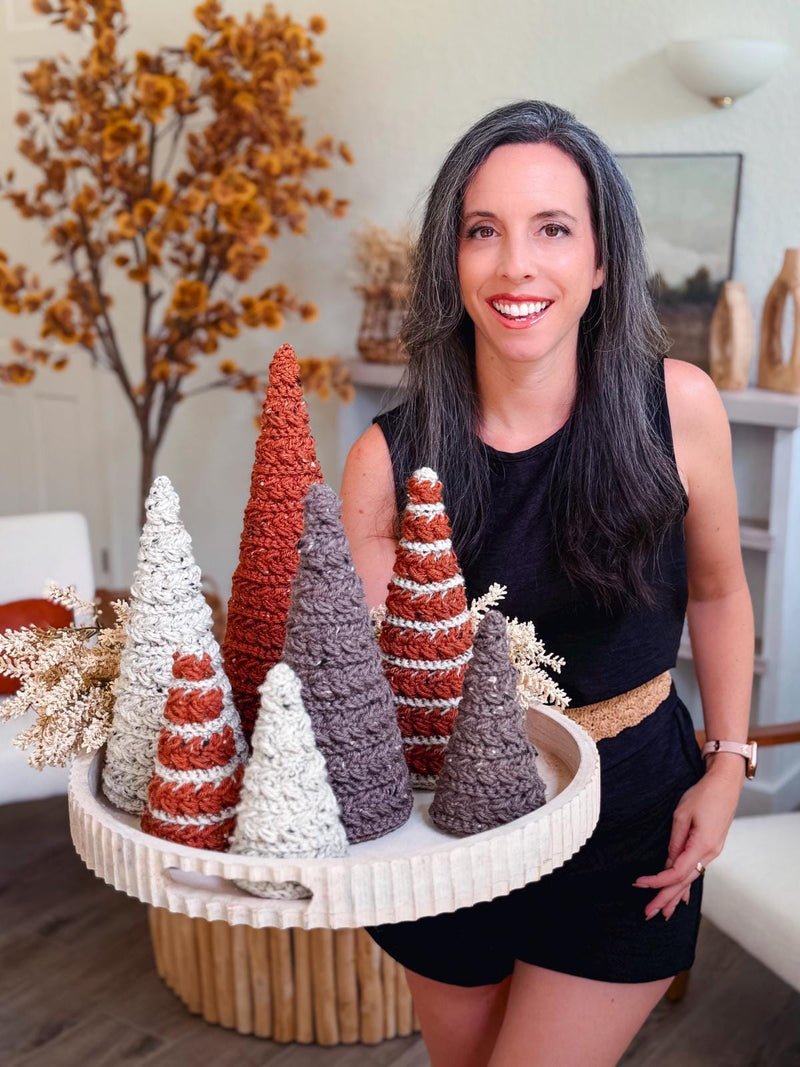Woman in black dress holding a round tray with decorative, crochet textured cone-shaped trees in white, brown, and rust—perfect as a handmade gift. Fall foliage and neutral decor accent the background for a cozy touch.