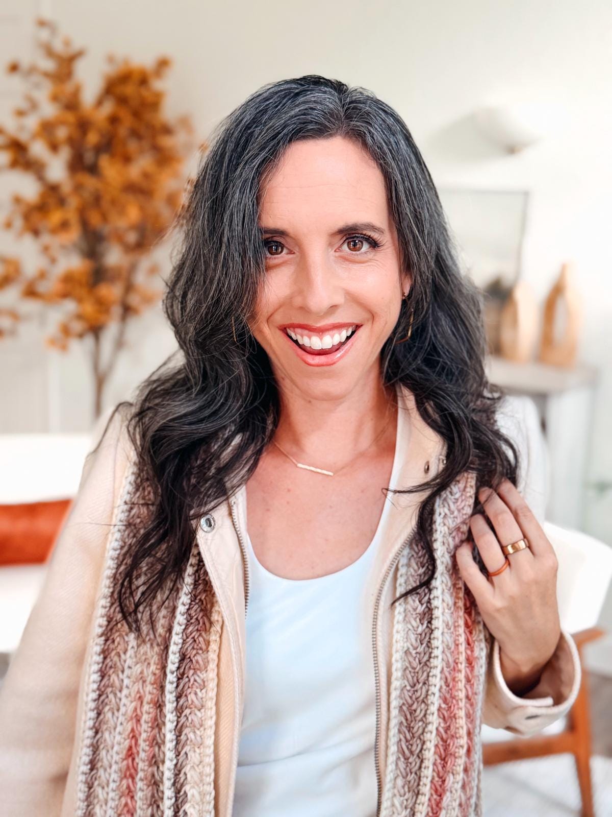 A woman with long dark hair and a streak of gray smiles at the camera, wearing a crochet scarf, light jacket, and white top—her style complemented by unique jewellery—in a bright room with autumn decor.