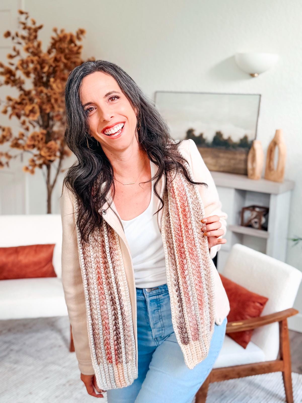 Woman with long dark hair, in a cream jacket, striped scarf, and jeans, stands smiling in a modern living room adorned with cozy autumn decor and soothing neutral tones.