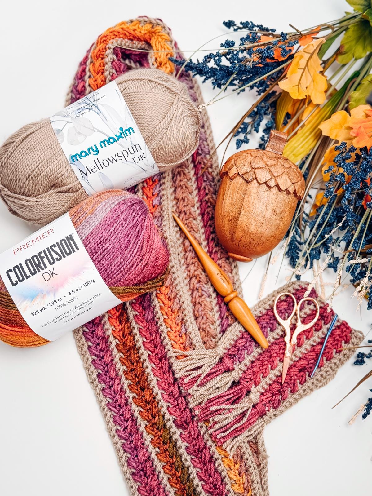 Yarn skeins, a crocheted scarf, crochet hook, scissors, a wooden acorn container, dried flowers, and unique jewellery are artfully arranged on a white surface.