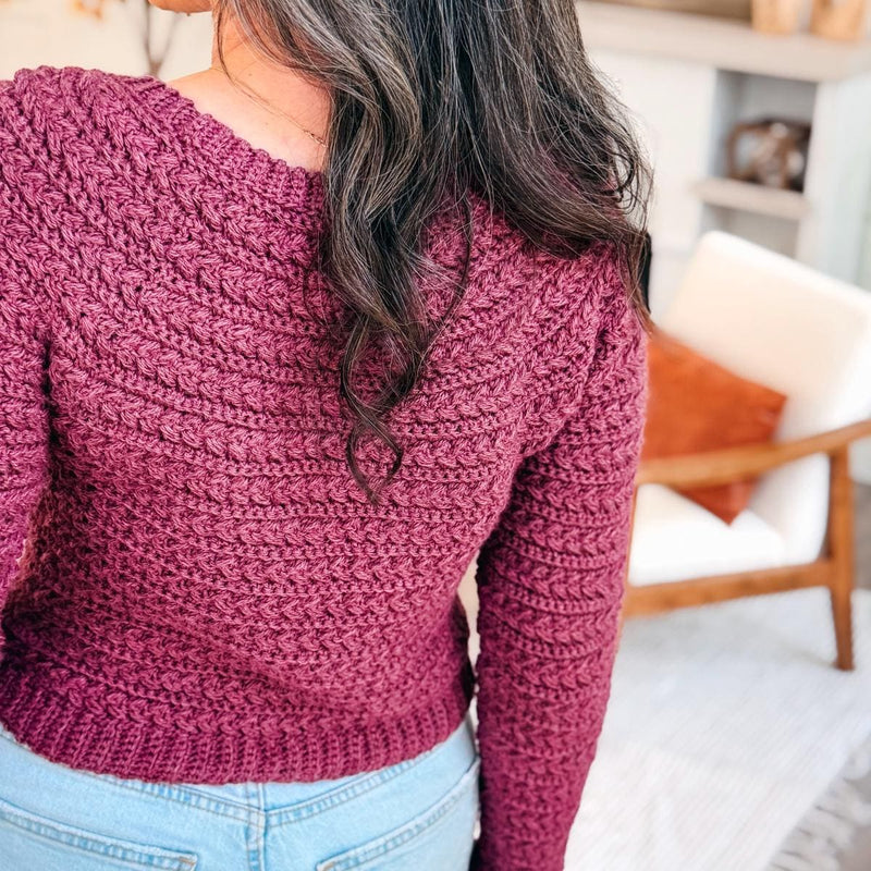 Back view of a plum crochet sweater with textured braided rows and ribbed hem worn with light jeans