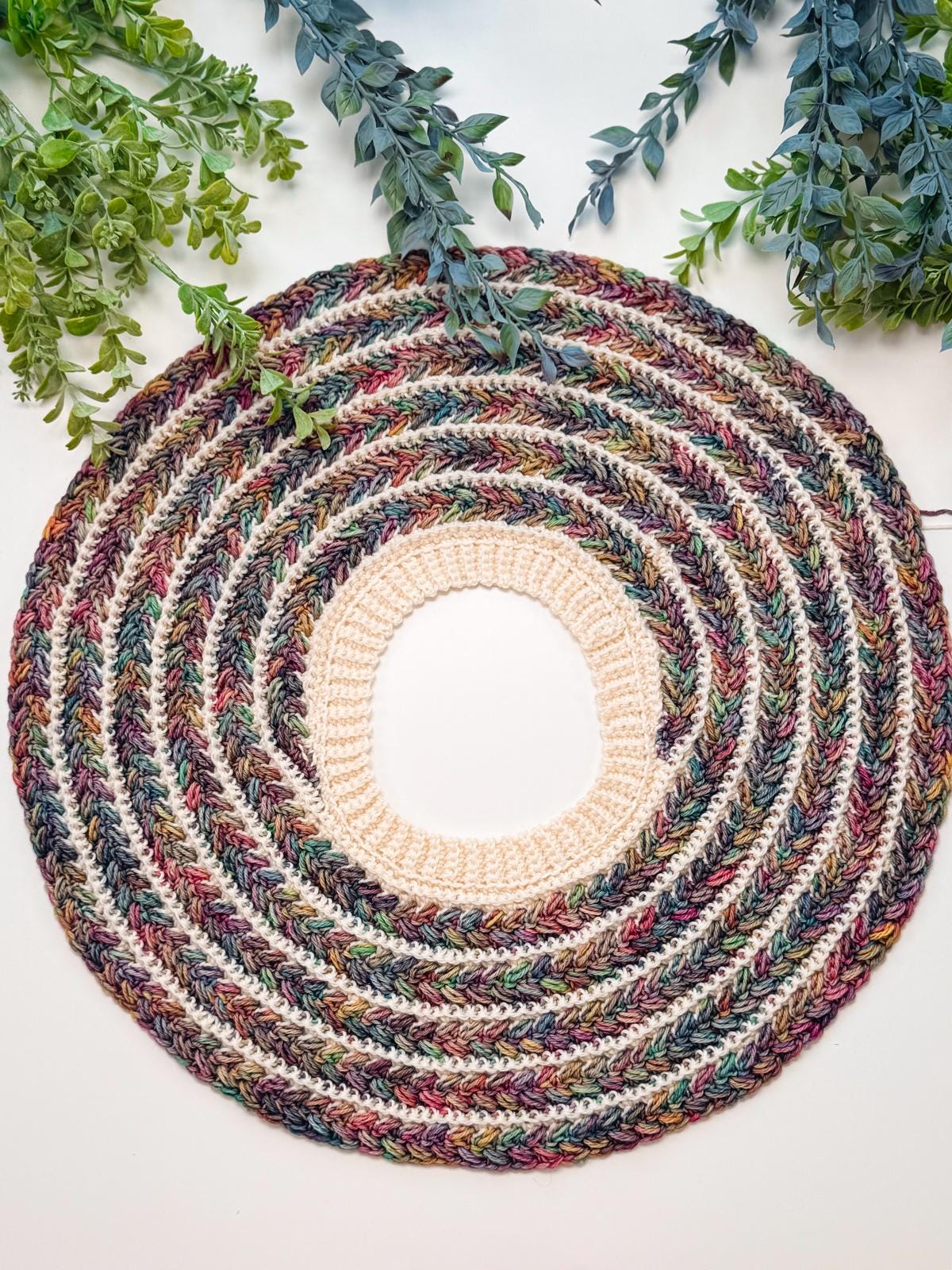 A round crocheted piece with a beige center and multicolored concentric rings, perfect as a handmade gift, is surrounded by green leafy branches on a white background.