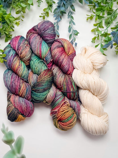 Three skeins of multicolor variegated yarn and one cream skein laid on white with green foliage above