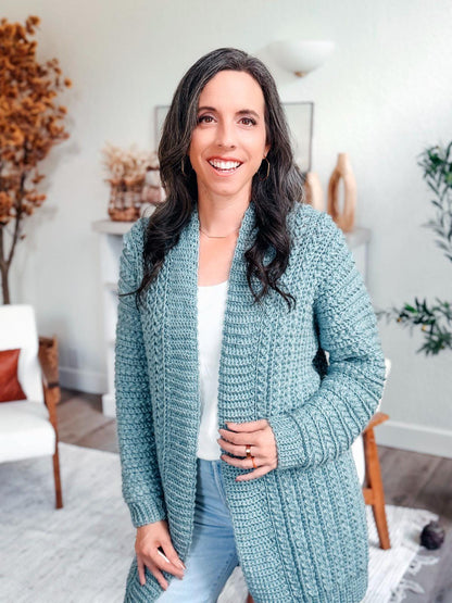 Sage blue crochet cardigan with textured sleeves and ribbed cuffs worn open by a smiling woman