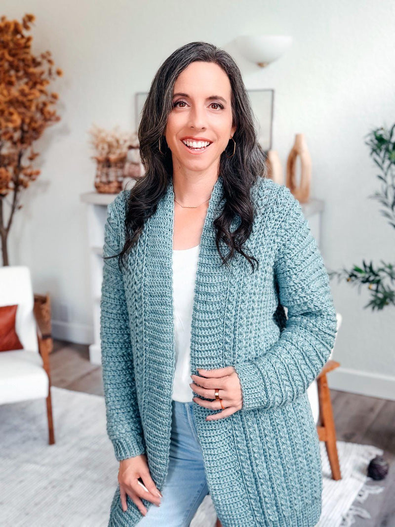 Sage blue crochet cardigan with textured sleeves and ribbed cuffs worn open by a smiling woman