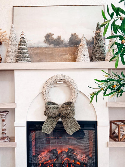 Cream crochet wreath with a green bow on a fireplace with crochet trees and a landscape painting above