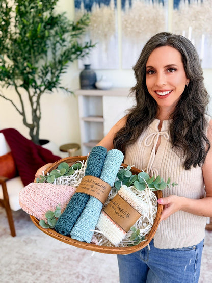 Woman in a cream top holding a basket of labeled crochet washcloths in pink, teal, aqua, and cream