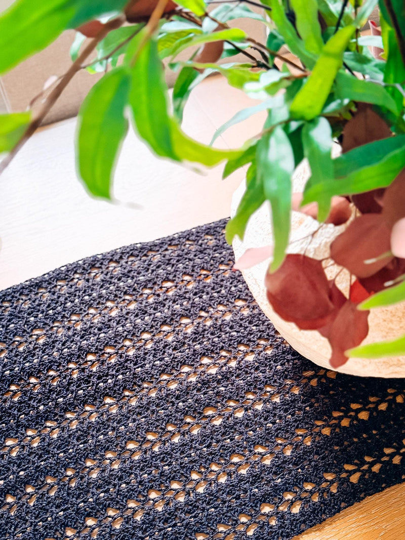 Close-up of a black crochet table runner showing its lacy mesh stitches beside a vase with green leaves