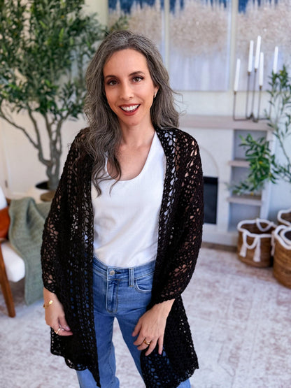 Black crochet mesh wrap worn open like a cardigan over a white top and jeans by a smiling woman indoors