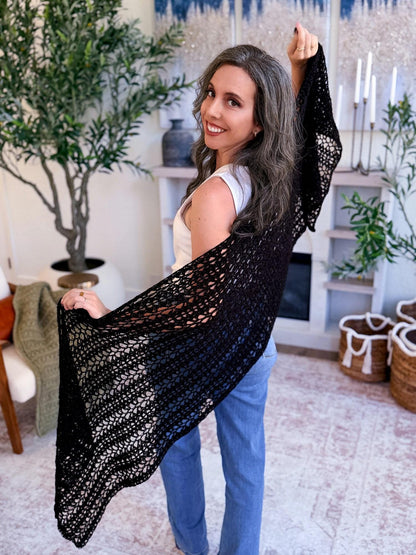 Black lacy crochet wrap stretched out by a smiling woman in a white top and jeans in a cozy living room