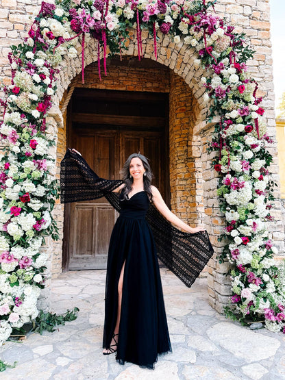Black crochet lace wrap held out by a woman in a black gown beneath a floral arch at a wooden door