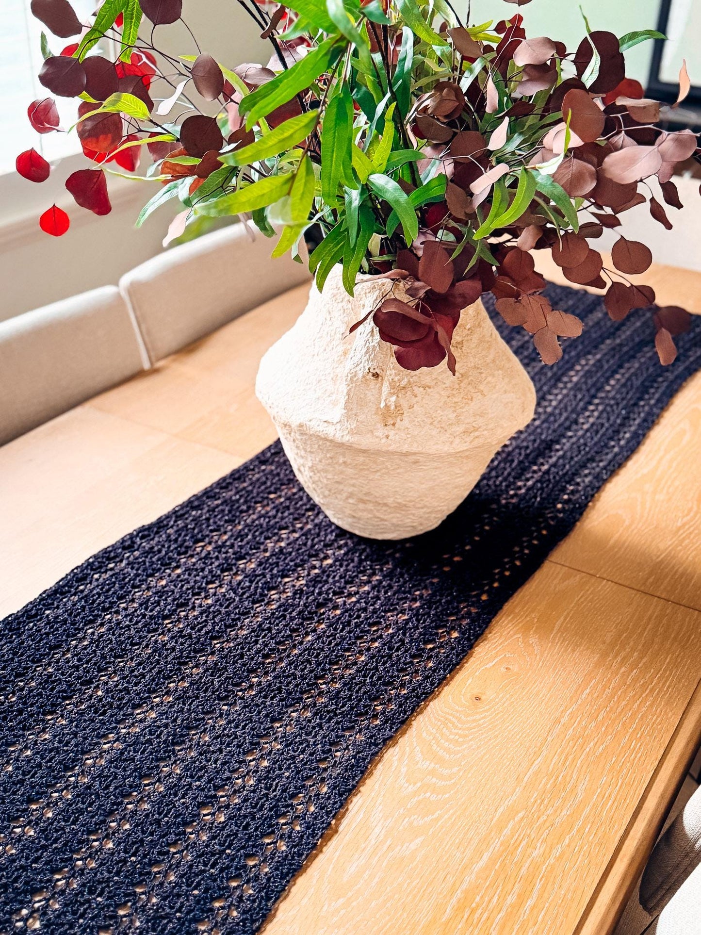 A cream-colored vase with red and green foliage sits on a wooden table with a dark woven table runner, creating the perfect setting for displaying unique jewellery or presenting a thoughtful handmade gift.