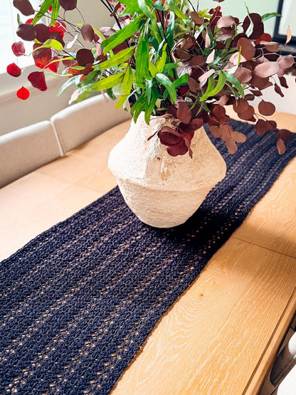 Black lacy crochet table runner on a wooden dining table with a cream vase of green and burgundy leaves