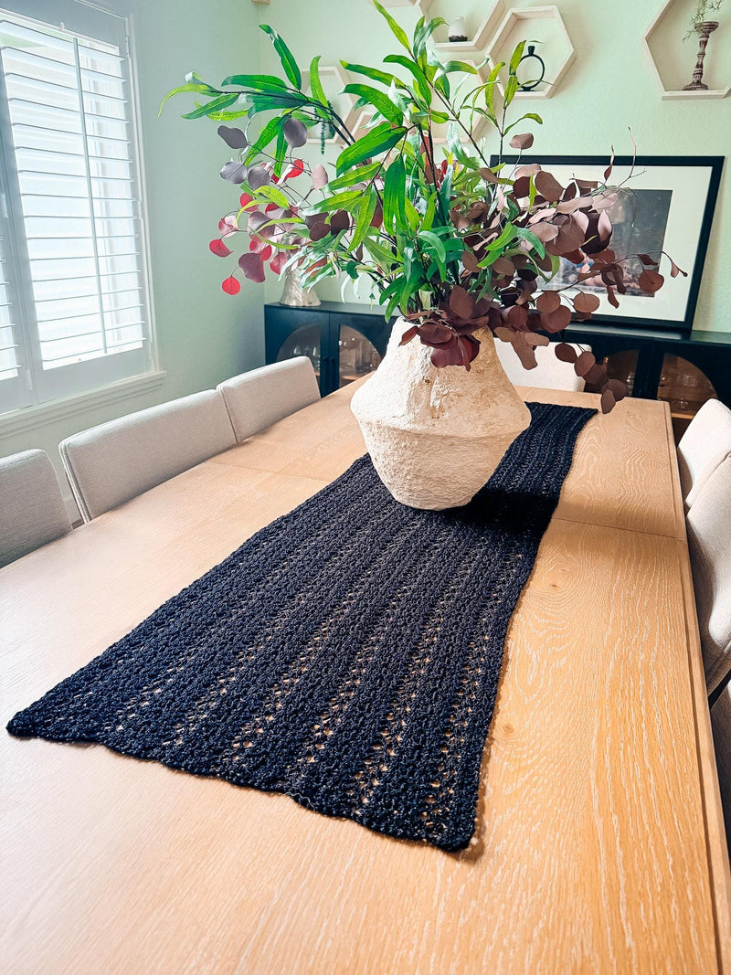 Black crochet table runner down a long wooden dining table with a cream vase of greenery in the center