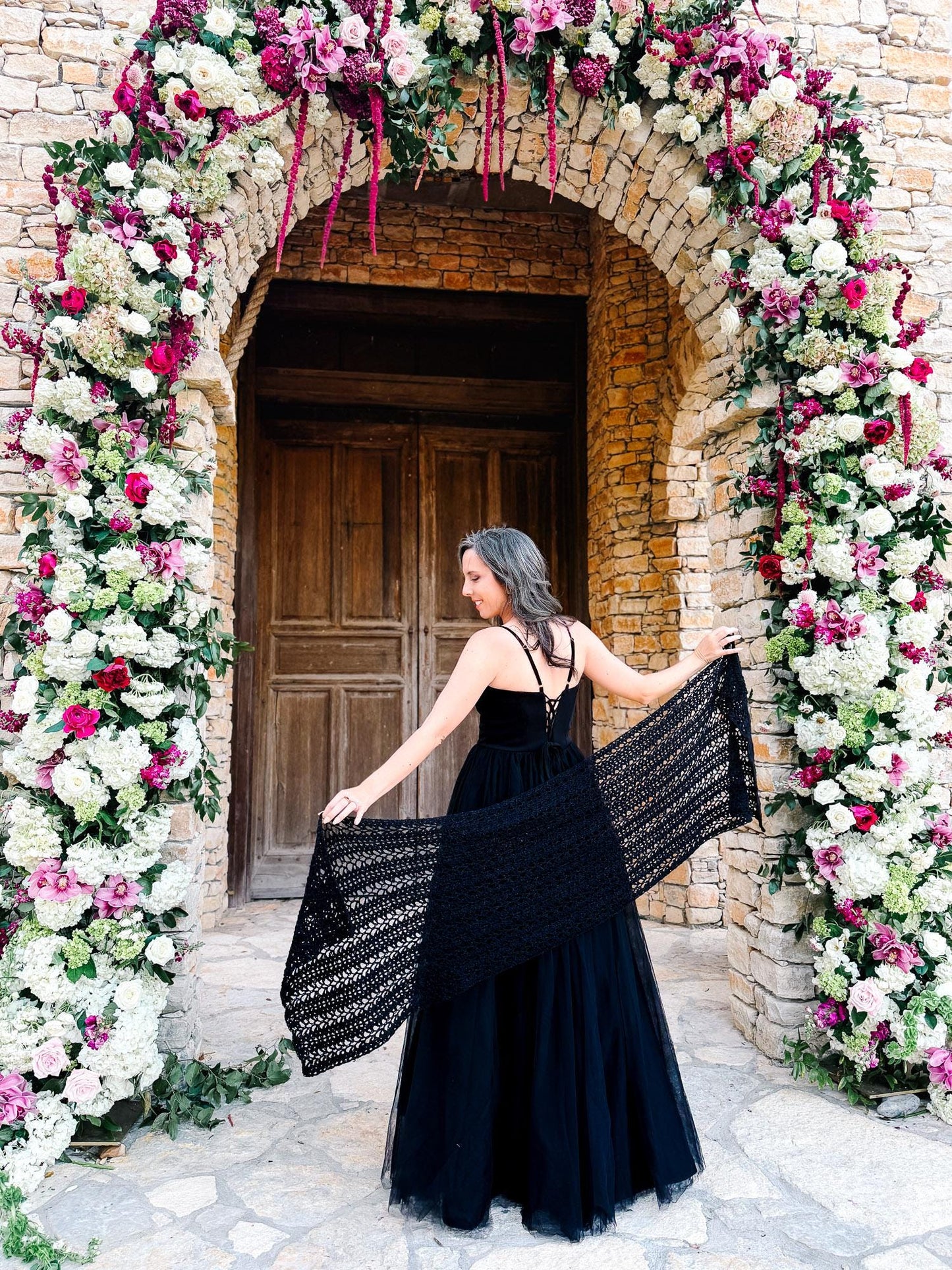 A woman in a black dress stands in front of a large wooden doorway framed by a floral arch made of white and pink flowers, holding out a black shawl behind her, perfectly accessorized with unique jewellery.