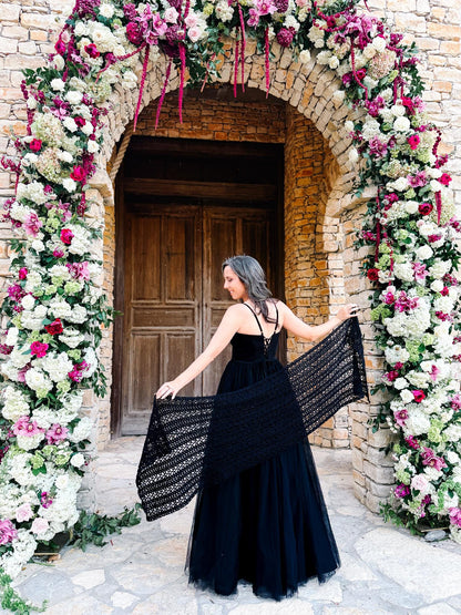 Black crochet mesh wrap held open behind a woman in a black gown under a pink and white floral stone archway