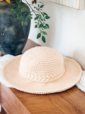 Cream crochet sun hat with braided band resting on a wooden table beside a dark vase and greenery