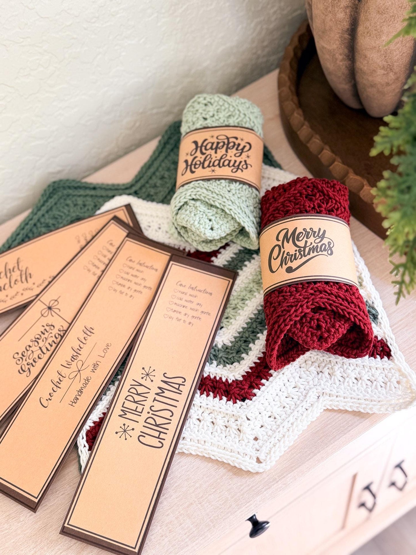 Two rolled crochet holiday washcloths in sage green and deep red are displayed on a light wood surface with decorative tags that read 'Happy Holidays' and 'Merry Christmas.'.