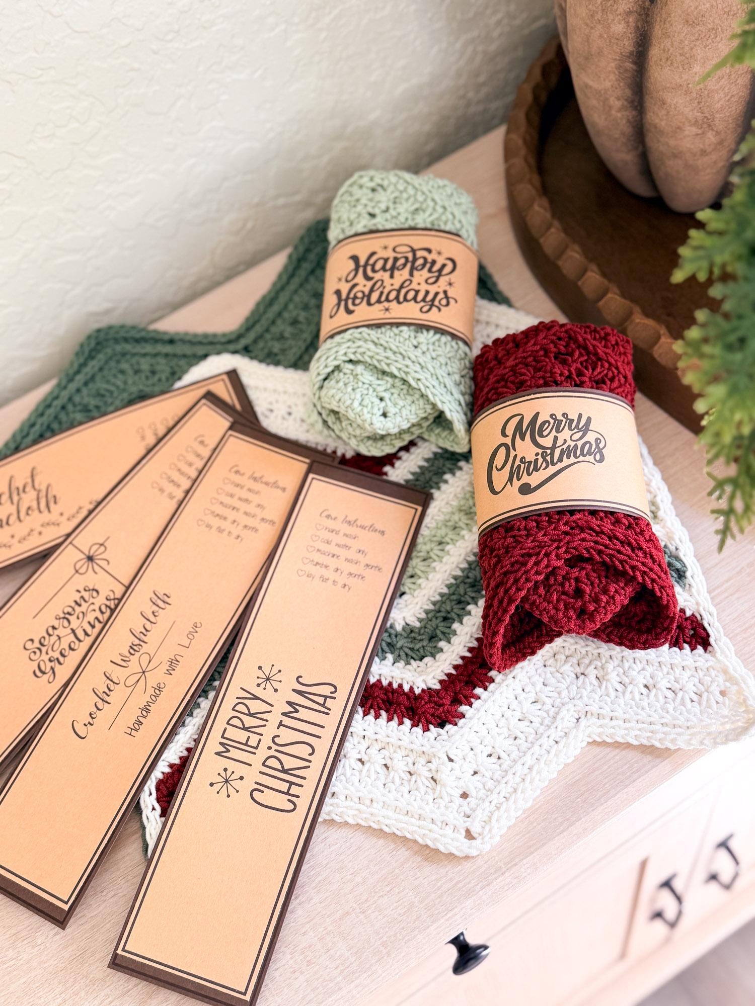 Two rolled crochet holiday washcloths in sage green and deep red are displayed on a light wood surface with decorative tags that read 'Happy Holidays' and 'Merry Christmas.'.