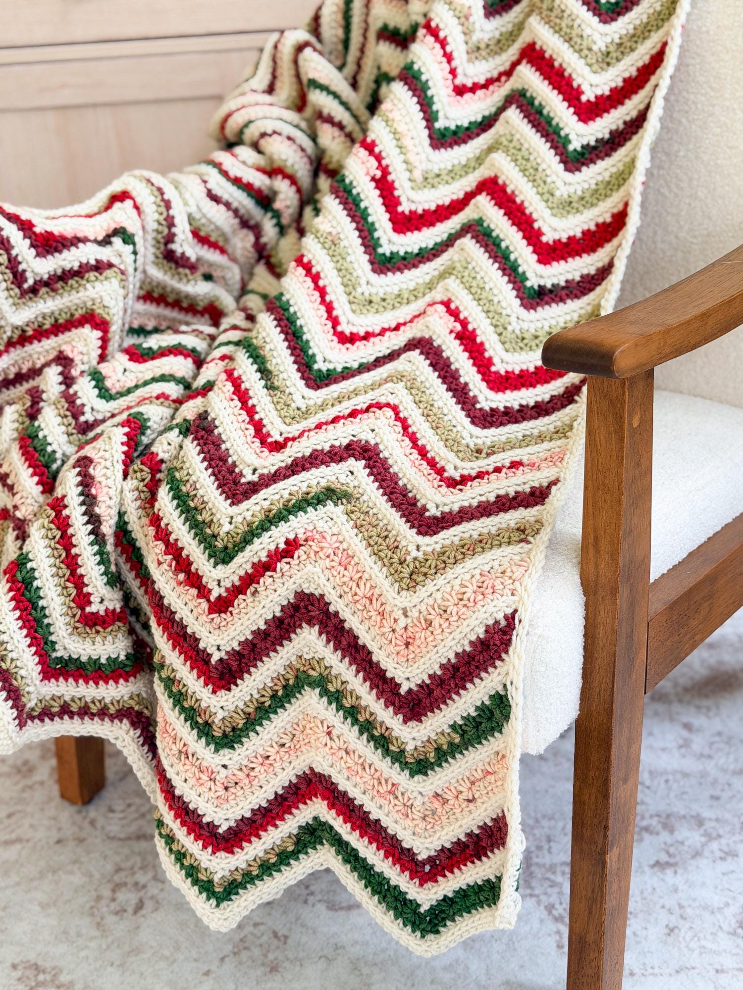 A chunky crocheted chevron throw blanket draped over a wooden armchair, featuring zigzag stripes of cream, red, green, and beige.
