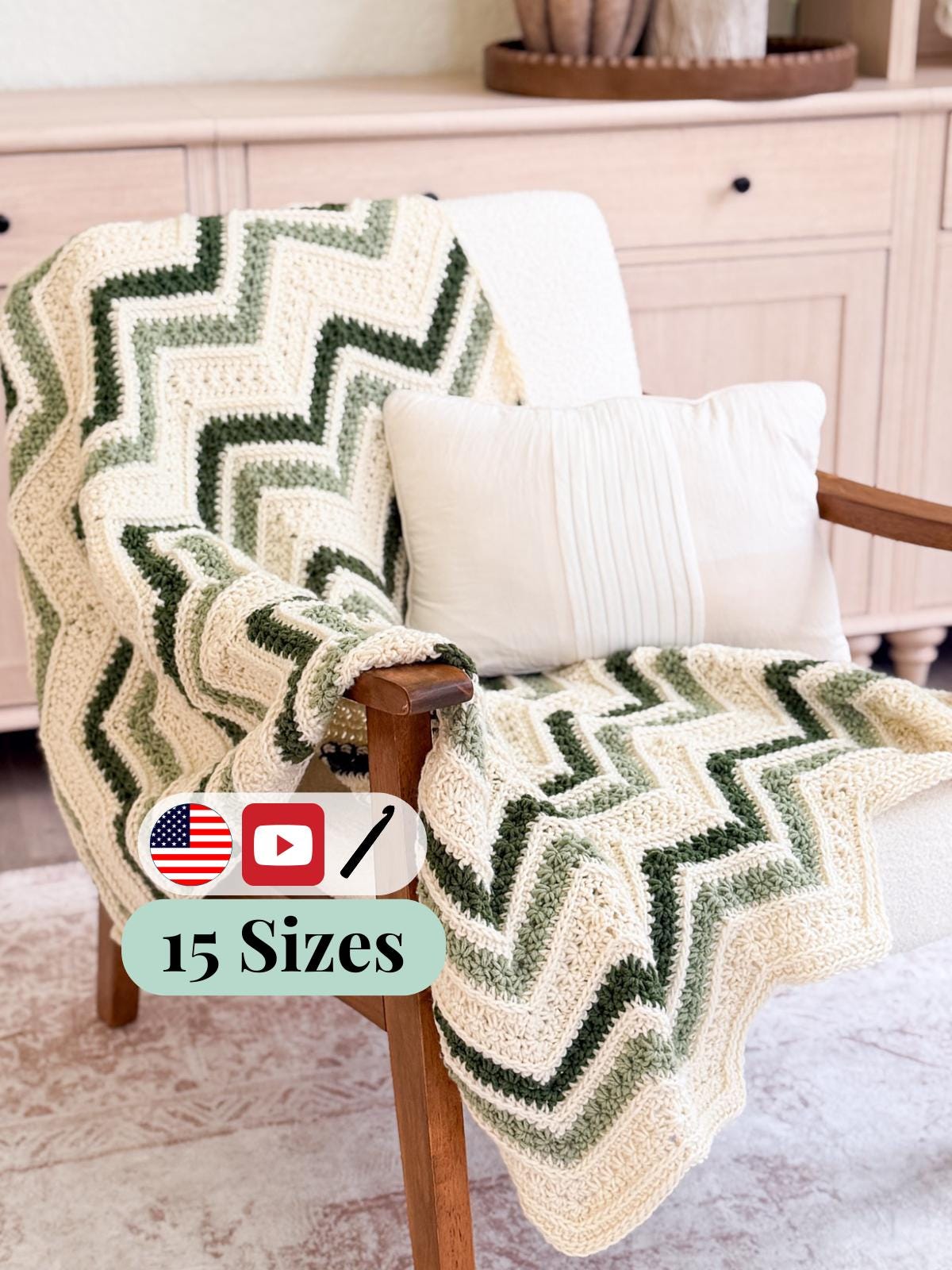 A crocheted chevron throw blanket draped over a wooden armchair, featuring alternating bands of cream, olive green, and dark forest green. A white pillow rests on the chair, with a light-colored cabinet in the background.