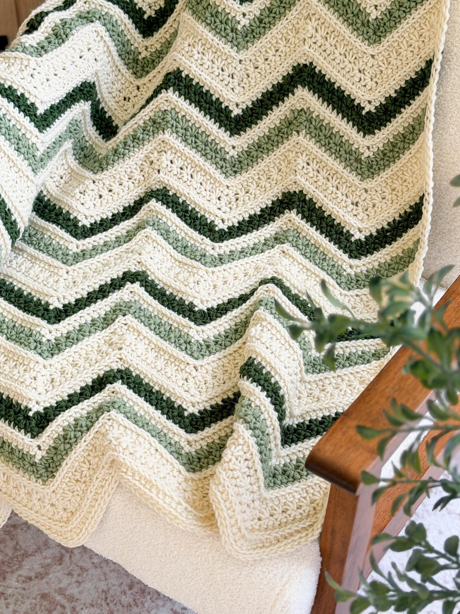 A crocheted chevron throw blanket draped over a chair, featuring alternating stripes of cream, sage green, and dark forest green.