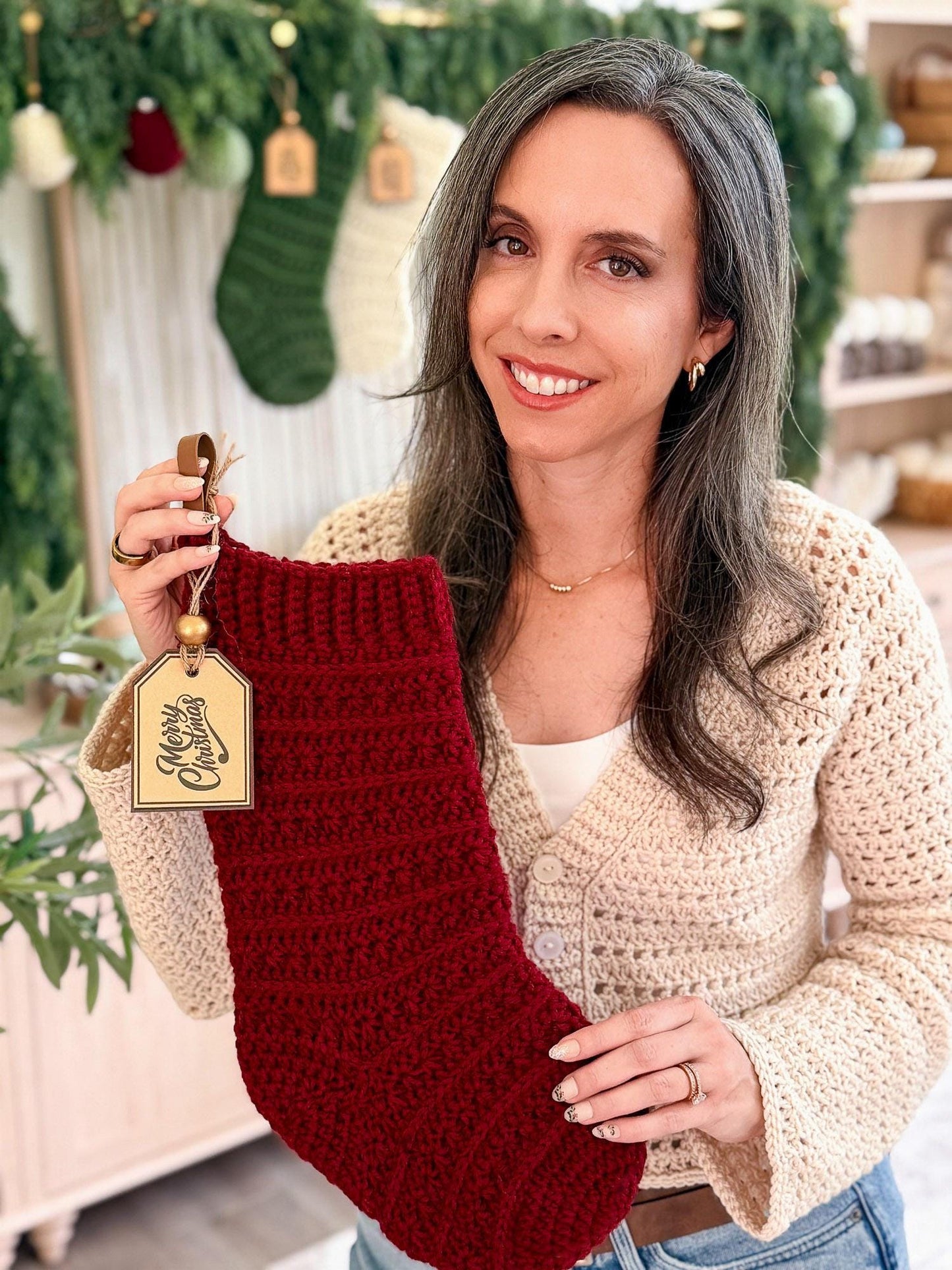 A smiling woman with gray hair holds up a dark red crochet Christmas stocking with a hanging tag. She is wearing a cream crochet cardigan in a decorated holiday shop setting with green garlands and other stockings in the background.