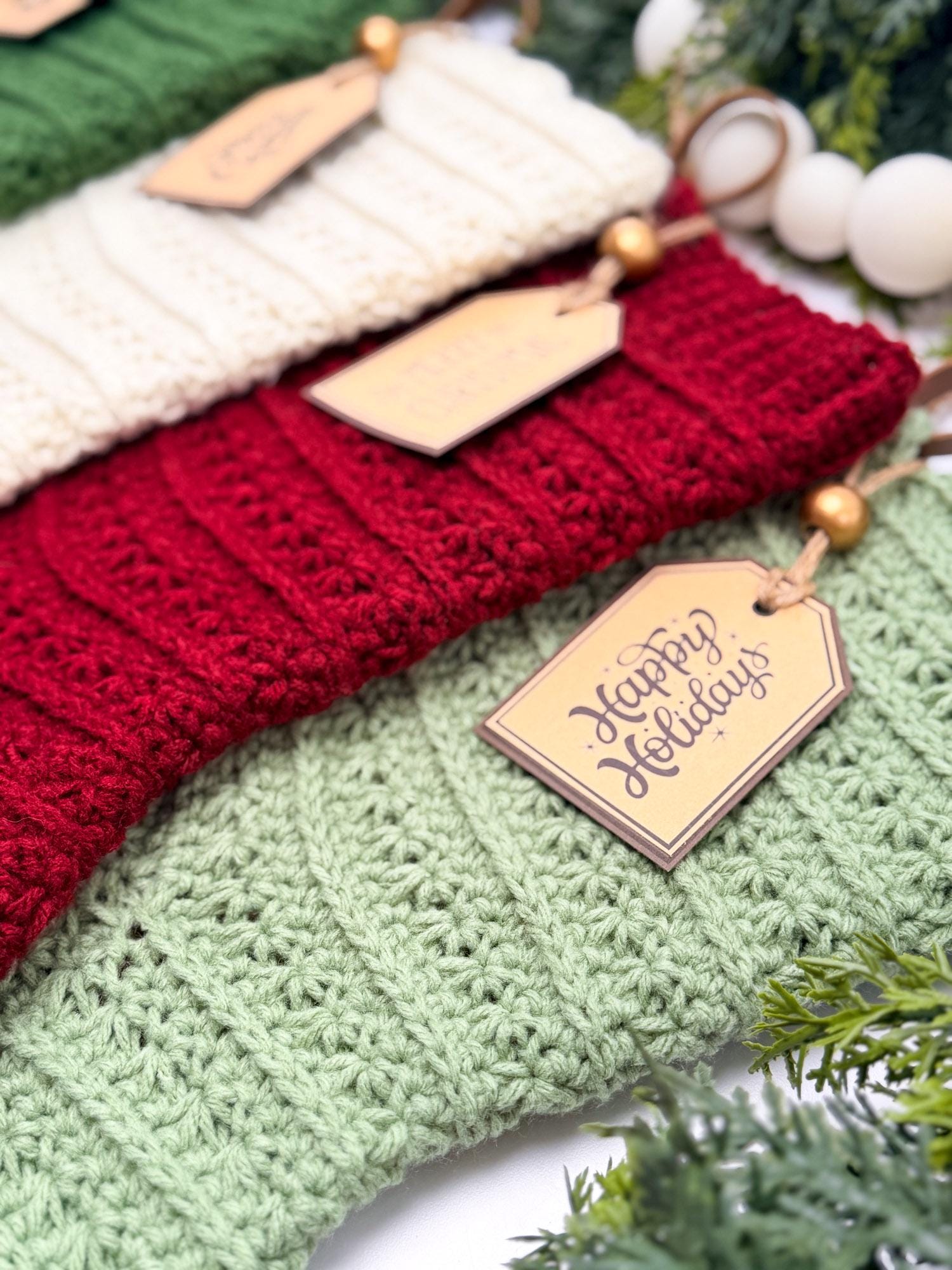 Three crocheted Christmas stockings in cream, red, and light green, each with a wooden gift tag and gold bead, arranged with holiday greenery and white beads.