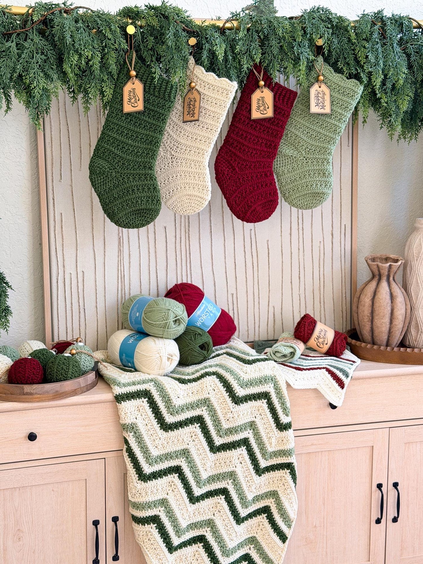 Four crocheted Christmas stockings in green, cream, burgundy, and sage hang from a garland above a light wood cabinet. A cream and green chevron-patterned blanket is draped over the cabinet, with balls of yarn and decorative vases nearby.