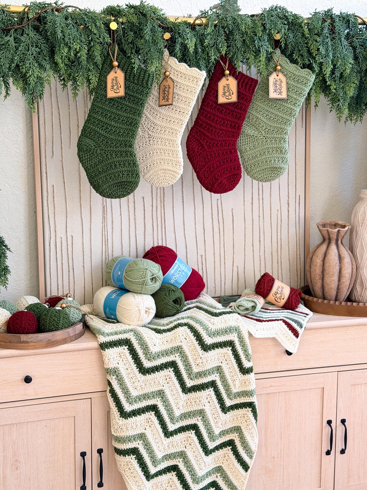 Four crocheted Christmas stockings in green, cream, burgundy, and sage hang from a garland above a light wood cabinet. A cream and green chevron-patterned blanket is draped over the cabinet, with balls of yarn and decorative vases nearby.