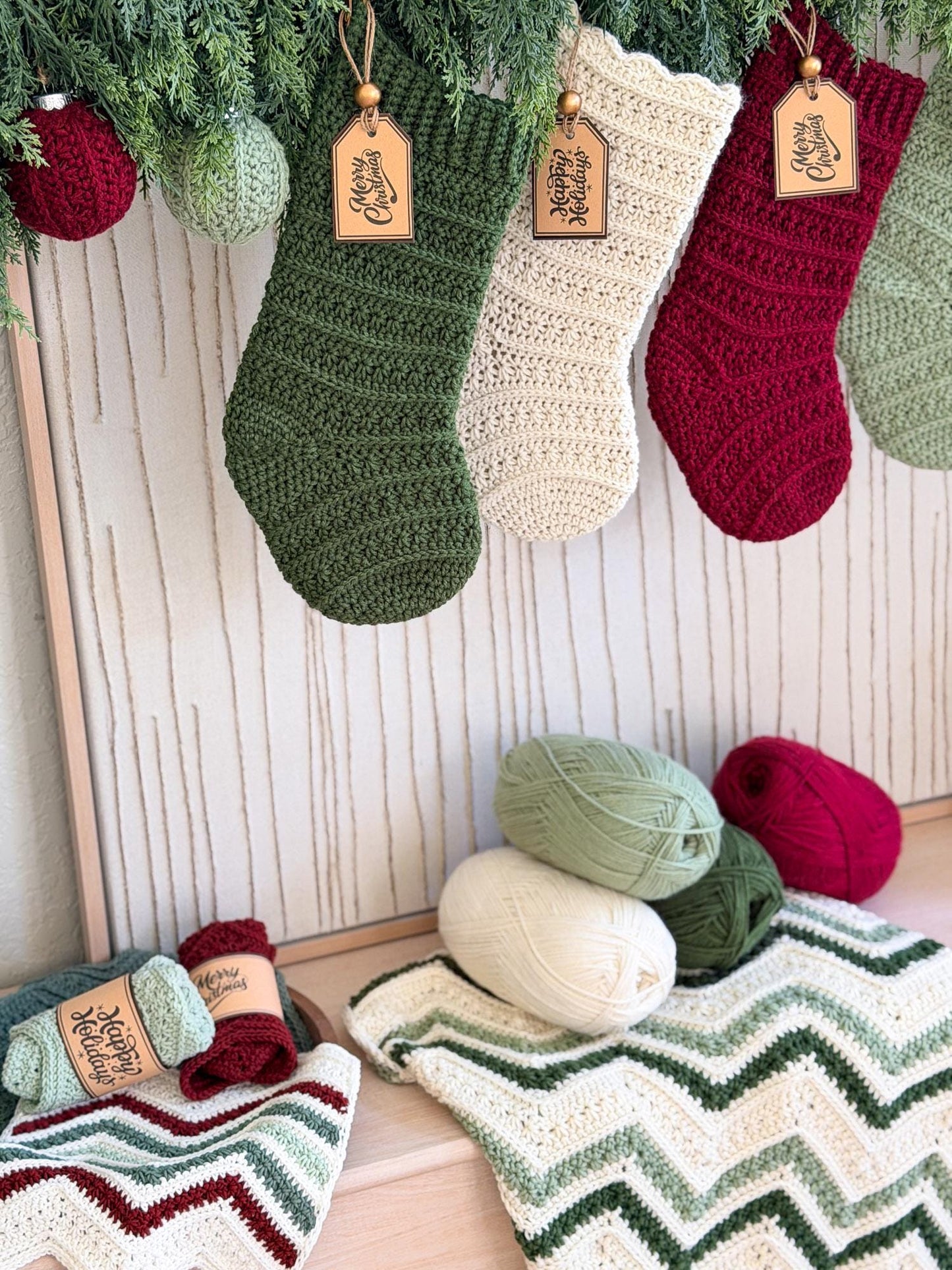 Three crocheted Christmas stockings in olive green, cream, and burgundy hang from a pine garland against a light wood paneled wall. Matching yarn balls and a chevron-patterned knit blanket are arranged below on a wooden surface.