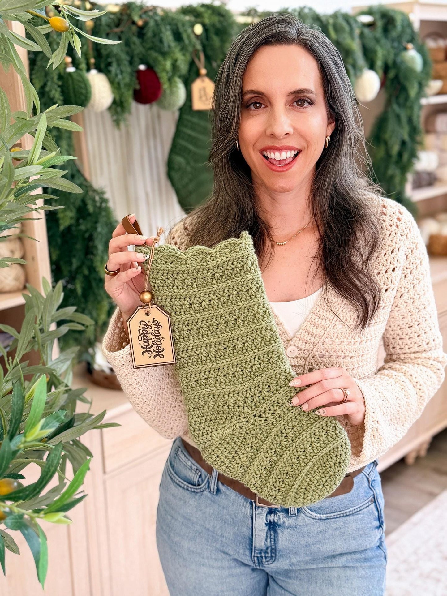 Woman holding a green crocheted stocking with a decorative tag in a festive setting.