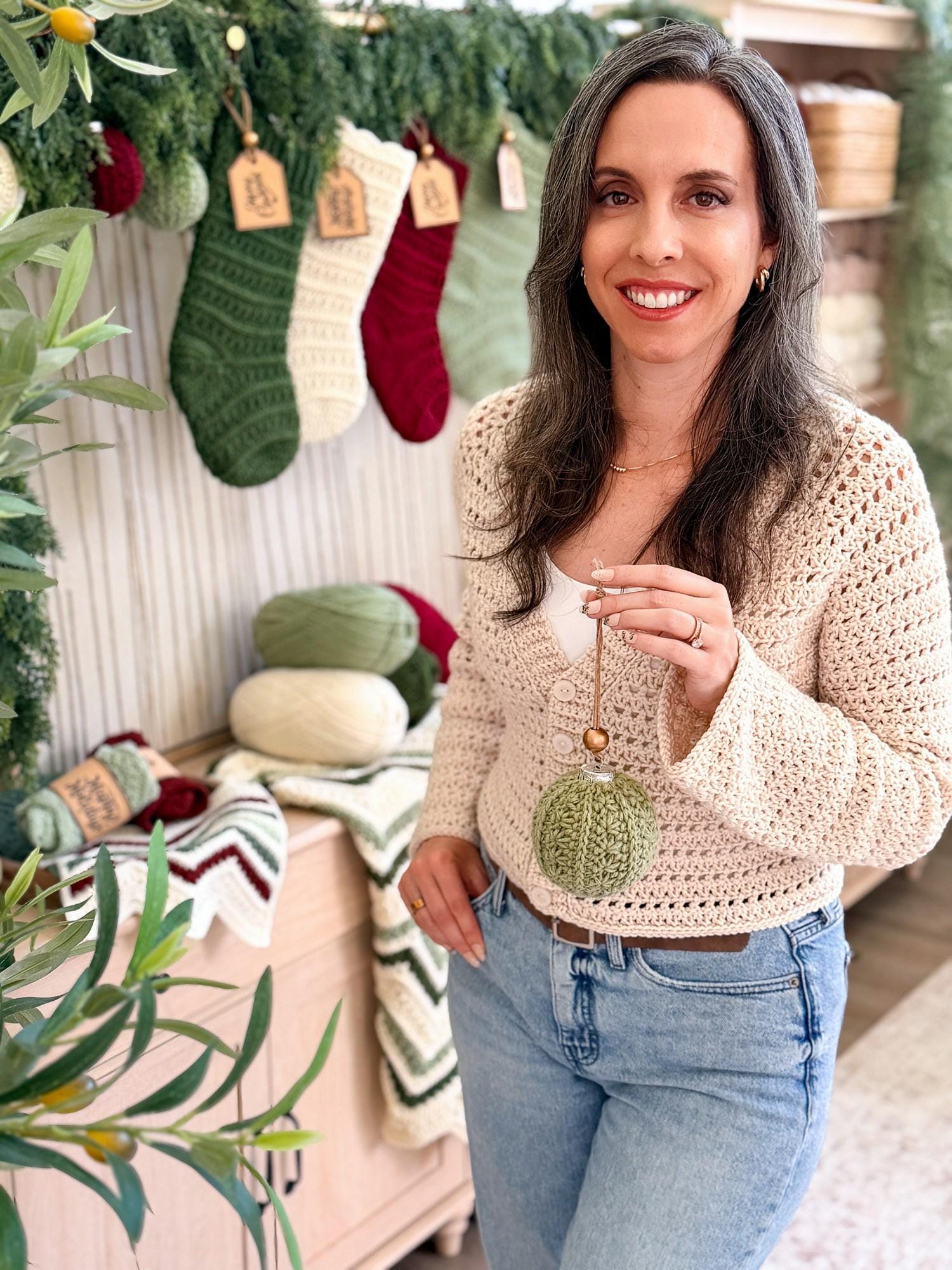 A woman wearing a cream-colored, long-sleeve crochet cardigan and light blue jeans, holding a green knitted ornament. The background features a cozy holiday display with green garlands, knitted stockings in green, cream, and red, and crochet blanket.